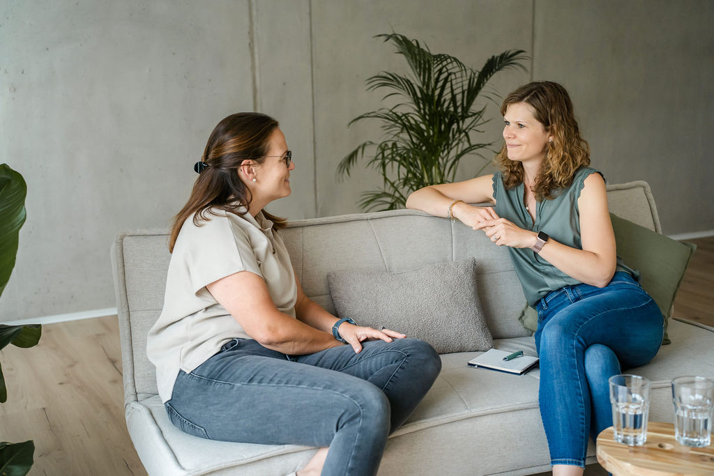 Coach mit Coachee im Gespräch auf einer Couch, Zettel und Stift liegen daneben.
